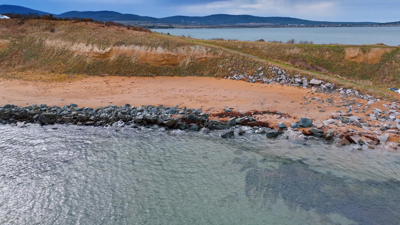 Spiaggia sabbiosa in riva all'acqua. La scena mostra una costa con spiaggia sabbiosa e zone rocciose. L'acqua calma riflette il cielo e le colline sullo sfondo