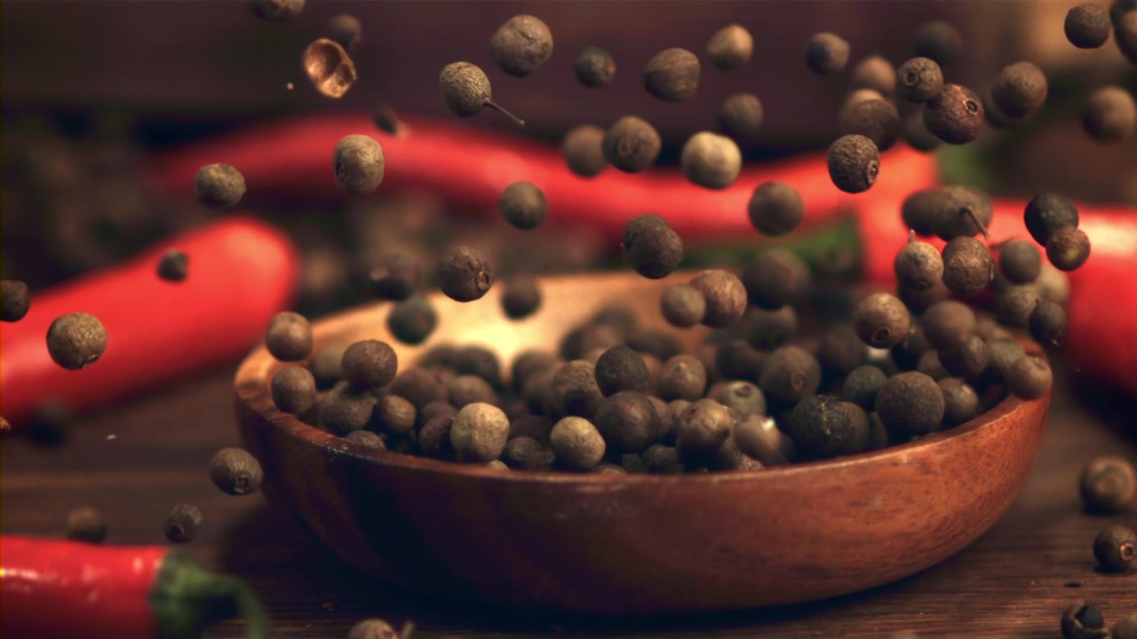 Granos de pimienta de Jamaica enteros cayendo en un cuenco de madera con chiles rojos.