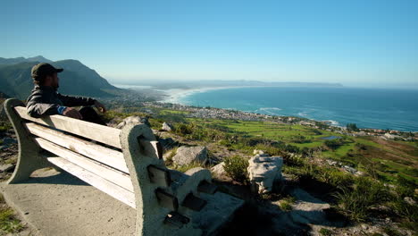 hombre sentado en un banco en el mirador de la montaña, con vista al pintoresco pueblo de pescadores, al campo de golf y al largo tramo de playa