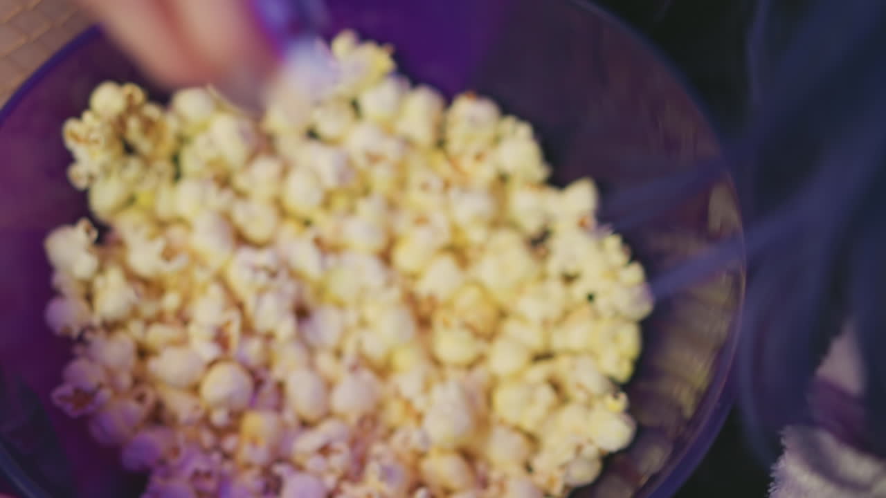 Close up of hand reaching into bowl filled with freshly popped popcorn under soft purple ambient light, capturing relaxed moment of casual snacking in cozy indoor