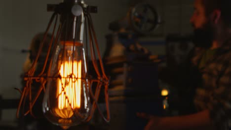 Closeup-of-a-work-light-and-then-a-worker-examining-a-piece-of-machinery-in-a-workshop