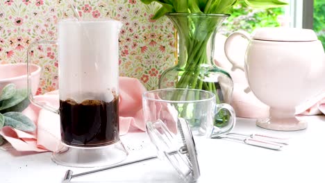 french press made of clear glass being used to make a hot coffee. pastel colors in a scene making hot coffee. fresh ground coffee for brewing using a french press method of extraction.