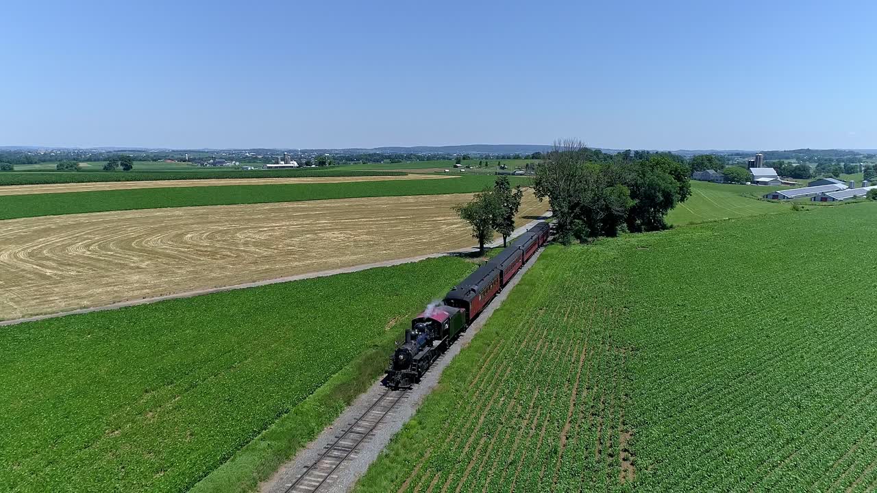 A steam train rolls through green fields and golden crops, showcasing the beauty of rural landscapes on a sunny day. Lush surroundings highlight the charm of vintage rail travel.