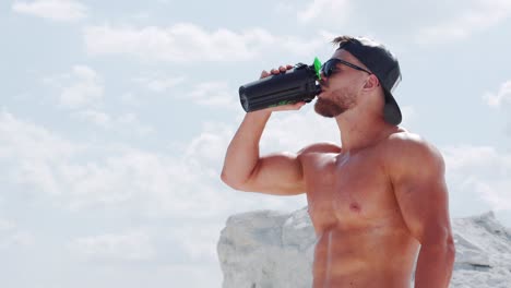 bodybuilder in a cap and sunglasses with large and strong muscles drinks water from a bottle. fitness exercise in nature