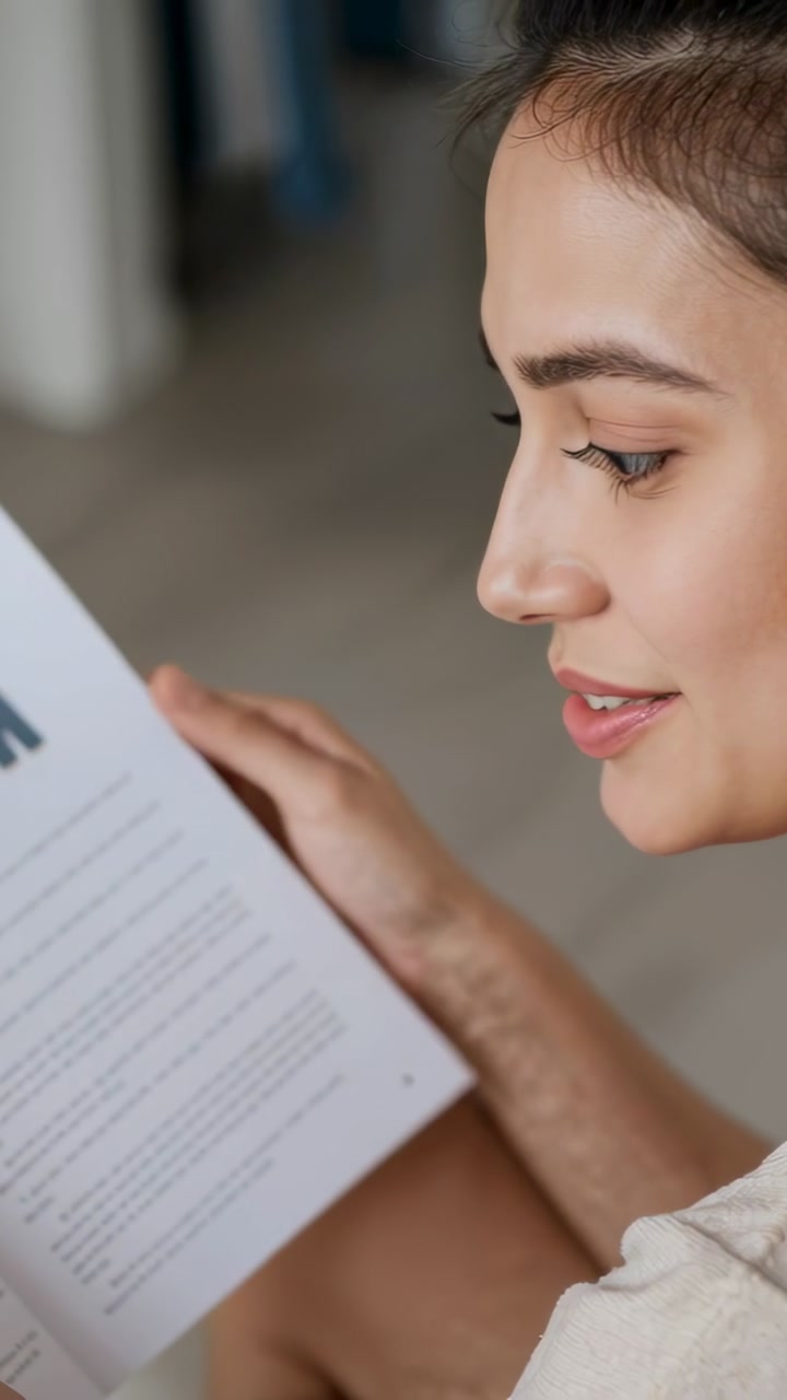 Glancing and smiling after camera prompt, woman turning booklet pages and reading at home