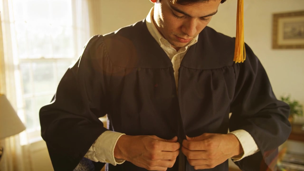 Manlig student knäpper och fäster mörk examenskåpa i varmt hem, gul tofs synlig.