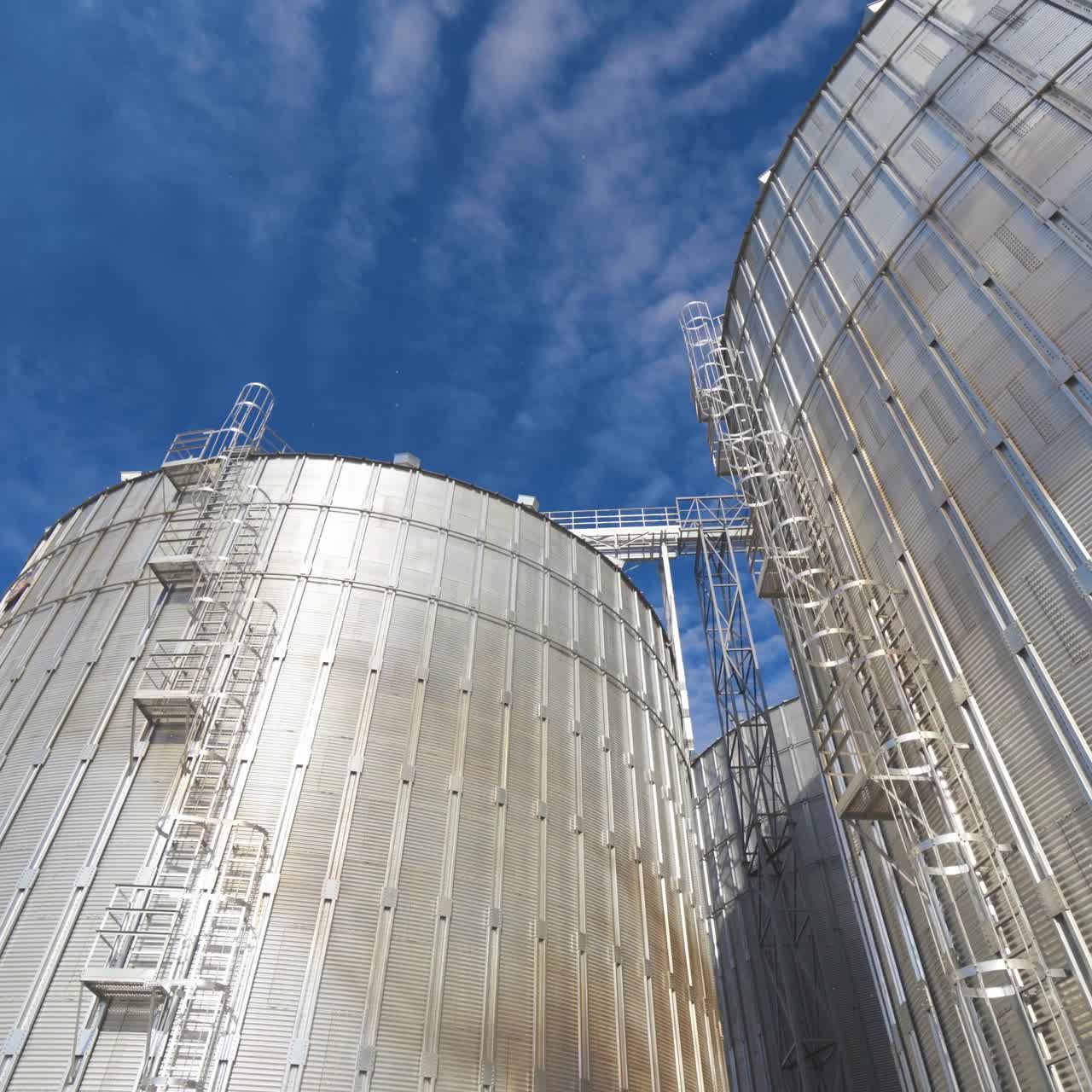 Silver storage tanks at sunlight. Large metal grain elevators against blue sky. Modern agro-processing factory