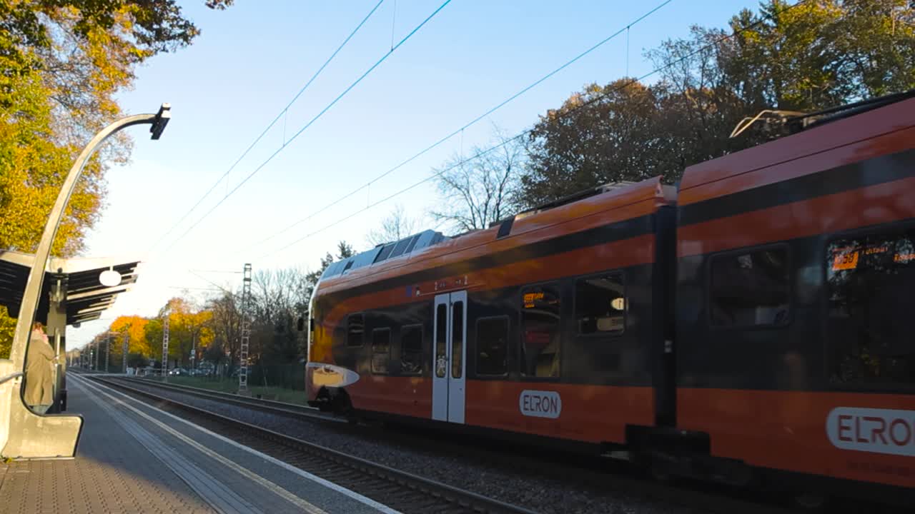 Orange public transport train that is powered by electricity or electric train passing by a train station or a railway station during autumn sunny day while trees around have yellow leaves, blue sky.