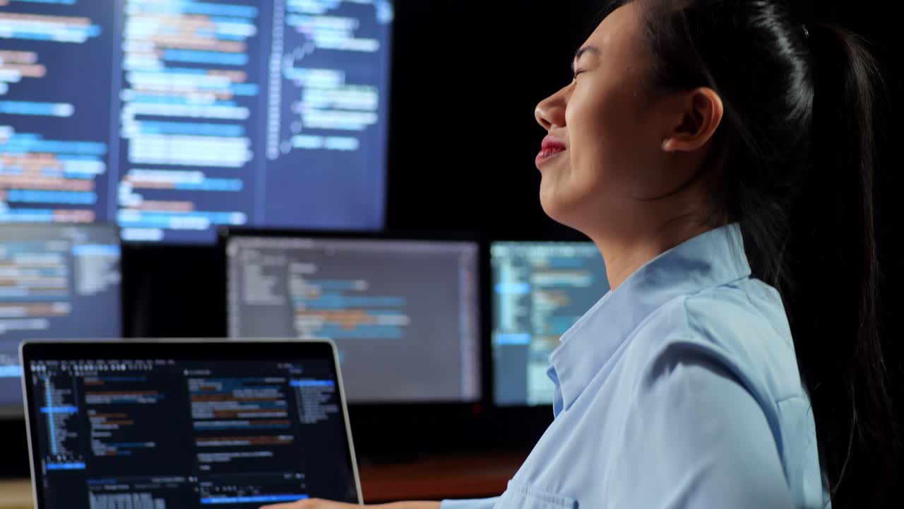 vista de cerca de una programadora asiática con dolor de espalda mientras escribe código en una computadora portátil usando múltiples monitores que muestran la base de datos en los escritorios de ventana de terminal en la oficina