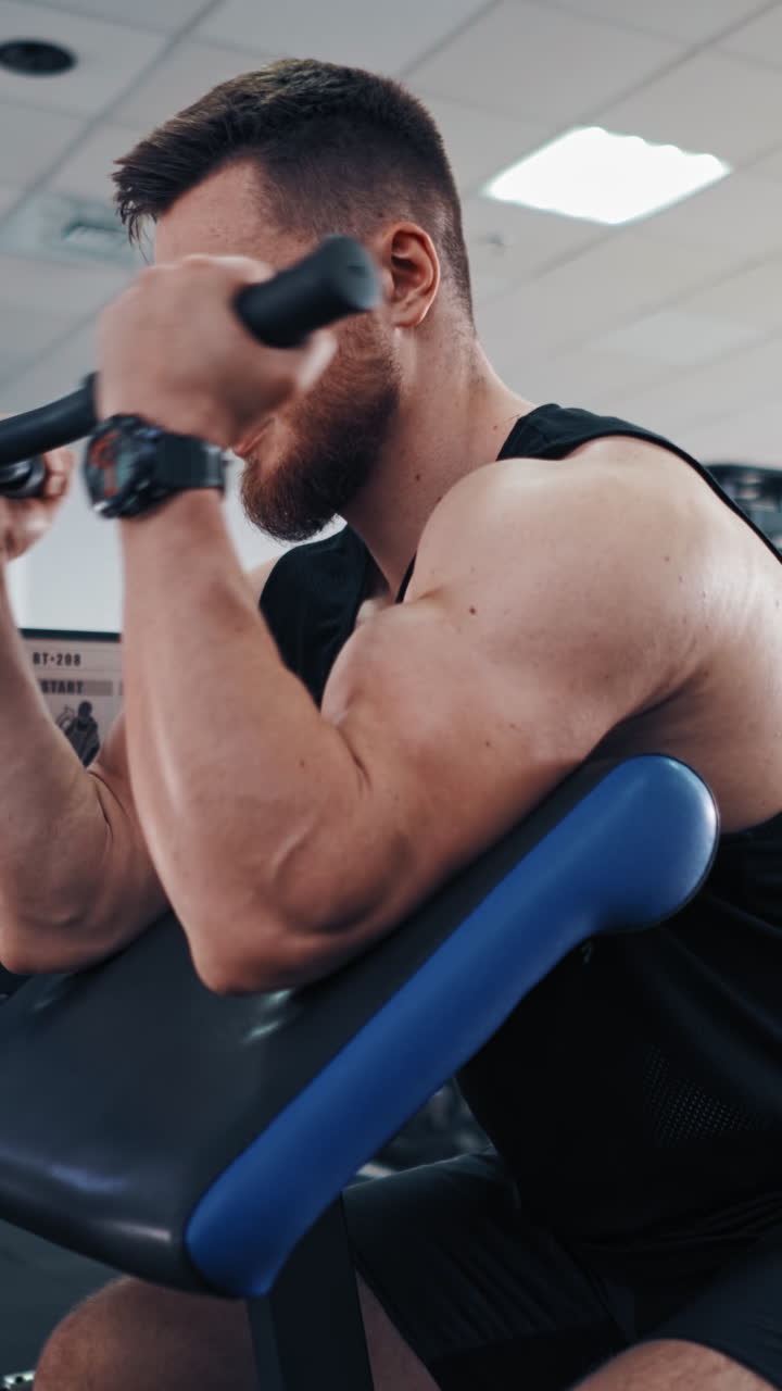Side view of an athletic man during workout in a gym. Bodybuilder sitting in a modern simulator and trains his muscles of arms. Vertical video