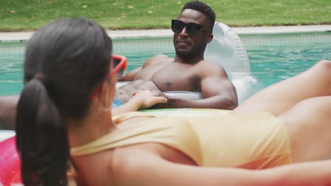 Happy-diverse-couple-wearing-swimming-suits-with-inflatables-at-swimming-pool-in-garden