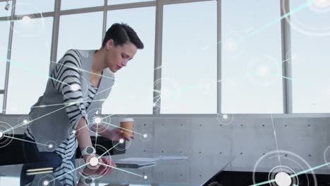 animation of network of connections over caucasian woman holding coffee cup reading a plan at office