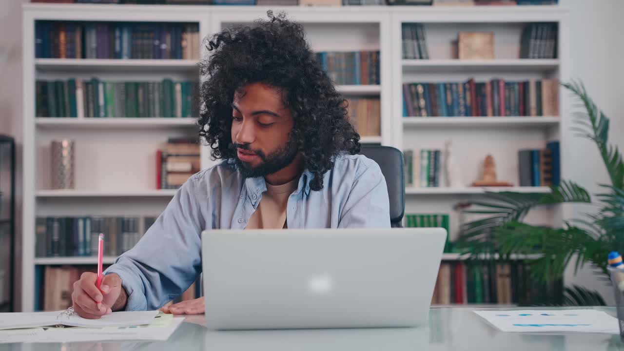 Focused arabian man student makes notes in notebook watching webinar on laptop