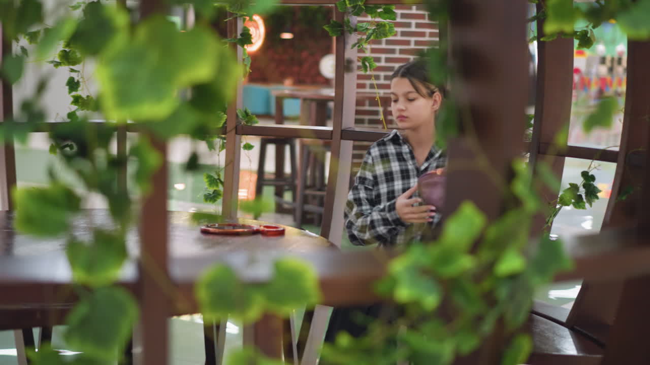 젊은 소녀는 현대적인 환경에서 아름답게 장식 된 의자에 앉아, 거울과 보라색과 파란색 지갑을 꺼내기 위해 학교 가방에 손을 고, 초록색 식물이 앉아있는 지역을 둘러싸고 있습니다.