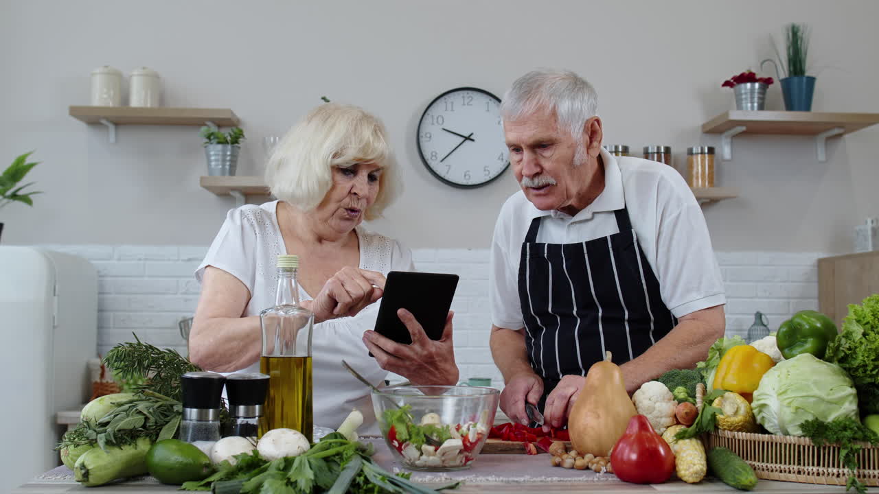 시니어 채식주의 커플이 생 채소로 러드를 요리하고 있습니다. 온라인 레시피를 위해 디지털 태블릿을 찾고 있습니다.