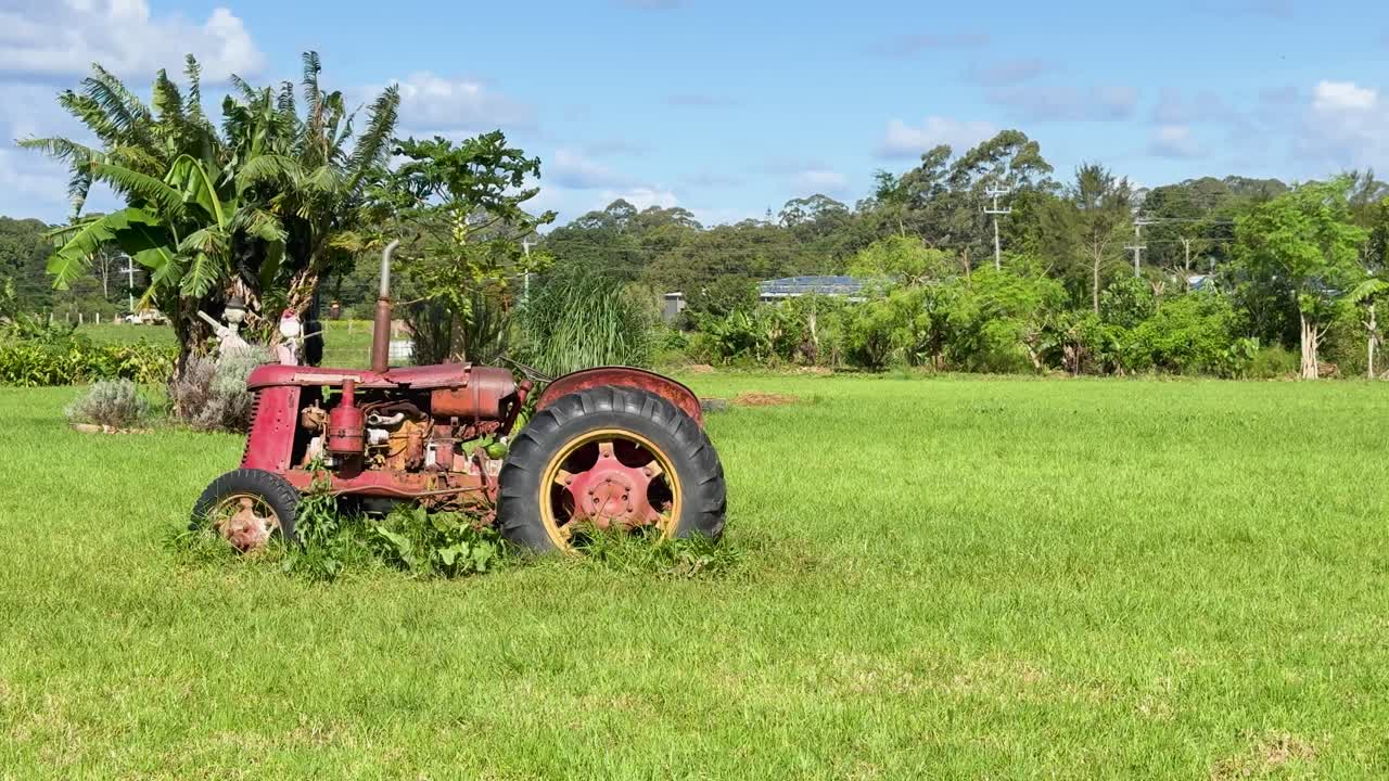 A stationary, weathered tractor sits in a lush green field under bright daylight, surrounded by overgrown grass and rural vegetation. Static camera, natural lighting
