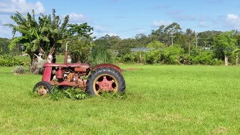 a stationary, weathered tractor sits in a lush green field under bright daylight, surrounded by overgrown grass and rural vegetation. static camera, natural lighting