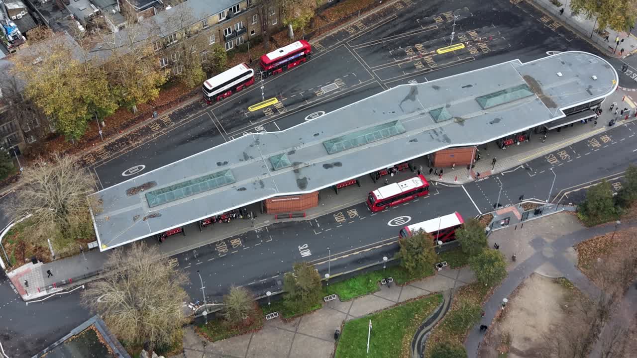 Static high-angle timelapse of Walthamstow bus station captured from above. Buses arrive and depart while passengers cross roads and board vehicles, showing urban transport hub activity in motion