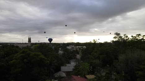 slow reveal of several hot air balloon rides taking off at dawn under a cloudy sky and a small town with native forest and a beautiful river on the outskirts