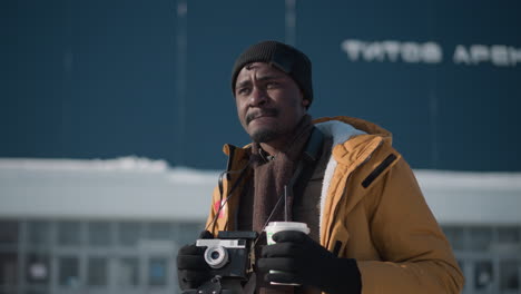 dark skinned videographer wearing winter coat and beanie holding camera and sipping drink while surveying urban scene under winter daylight, blurred cityscape background shows buildings lines