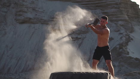 muscle athlete strongman man hits a hammer on a huge wheel in the sandy mountains in slow motion at sunset. the dust from the wheels rises.