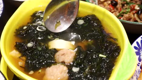 A spoon scoops silken tofu from a vibrant bowl of pork meatball and seaweed soup, under bright indoor lighting, with close-up camera movement