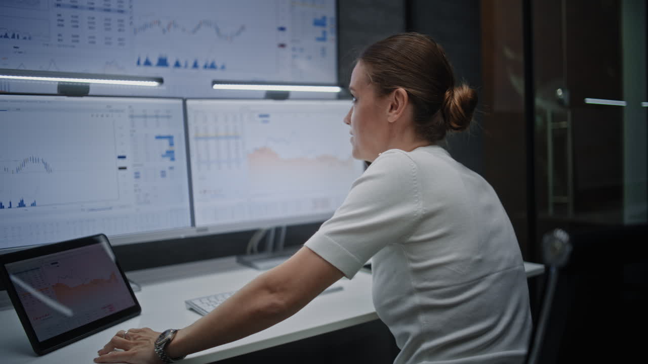 Female Financial Analyst Having Phone Call, Analyzing Real-Time Stocks on Multi-Monitor Computer and Digital Tablet. Businesswoman Working in Financial Agency Office at Night. Vertical Shot.