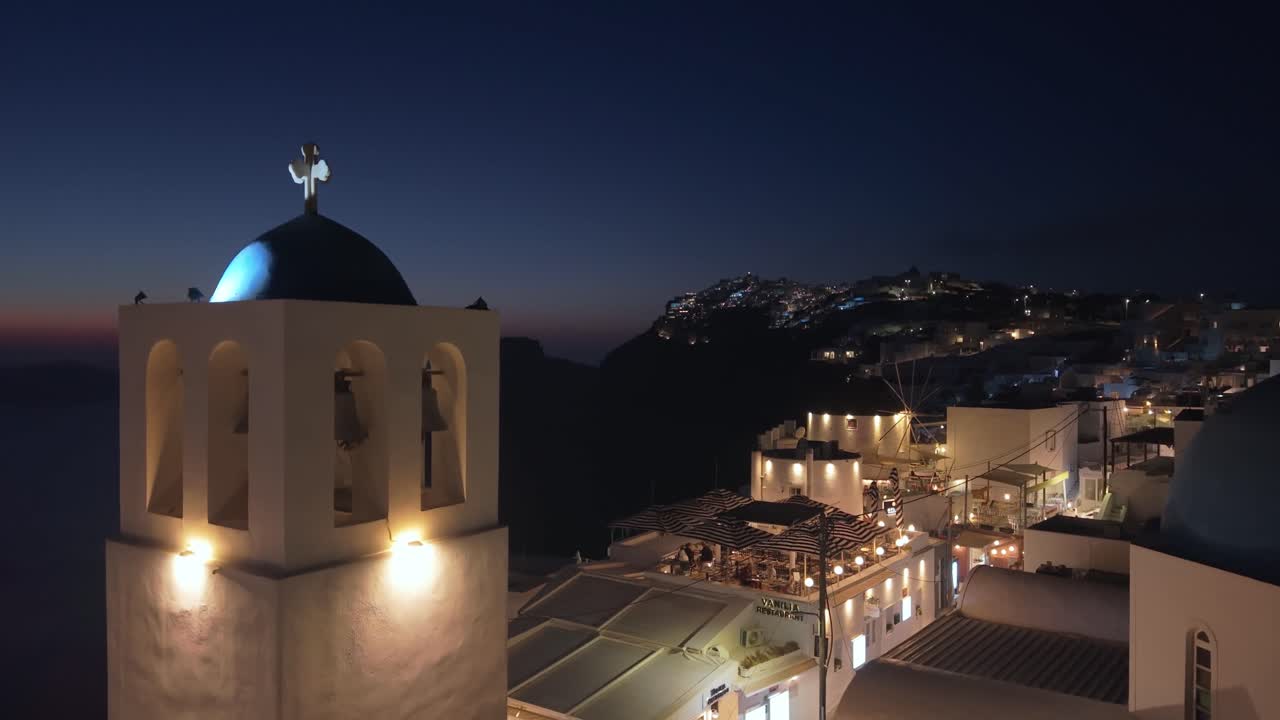 Una panoramica con drone al crepuscolo che inizia sopra le case illuminate di Fira, muovendosi lungo la caldera per rivelare la silhouette di Skaros Rock e il villaggio luminoso di Imerovigli all'orizzonte.