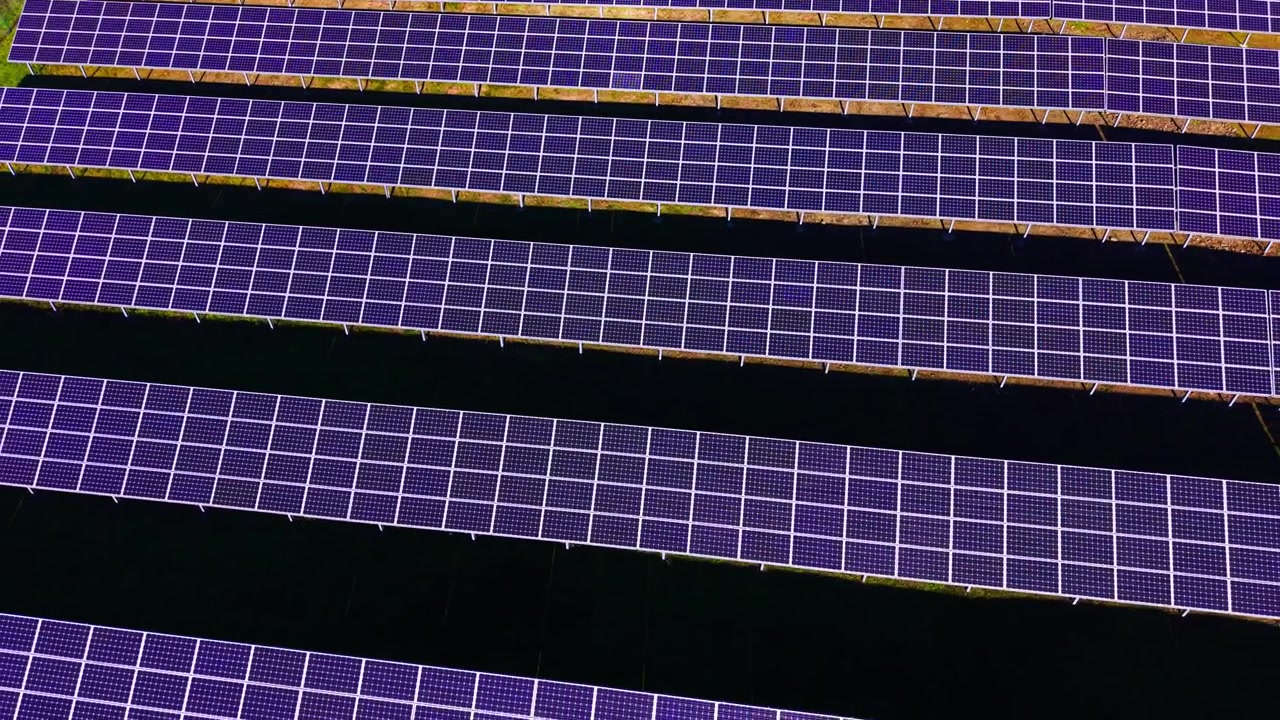 Vista drone di file di pannelli solari con sfumature viola. Linee diagonali di moduli fotovoltaici creano un motivo ripetuto su un terreno verde