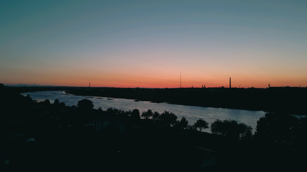 Cityscape captured at twilight with a river flowing under an orange sky, silhouetted trees and distant industrial structures creating a mysterious atmosphere