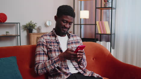 joyful young adult man sitting on sofa using smartphone watching video, online shopping, tapping