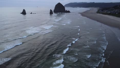 slow reveal shot of waves crashing onto cannon beach, revealing haystack rock in the background. the sunset on the oregon coast shows the forest, beach, and houses contrasting the sea stacks in 4k.