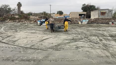 Los-Pescadores-Con-Botas-Amarillas-Llevan-Un-Bote-De-Remos-De-Fondo-Plano-Hasta-La-Orilla-A-Través-De-La-Playa