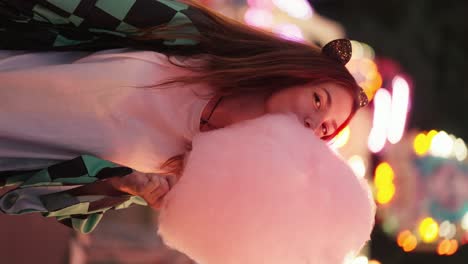 vertical video: portrait of a happy brunette girl with cat paws who bites and tears off a piece of large pink cotton candy during her walk in a bright amusement park in the evening