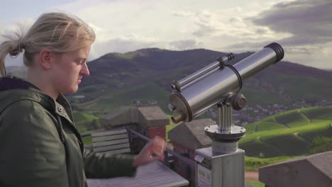 a blonde girl in a green raincoat looking through a telescope to the valley