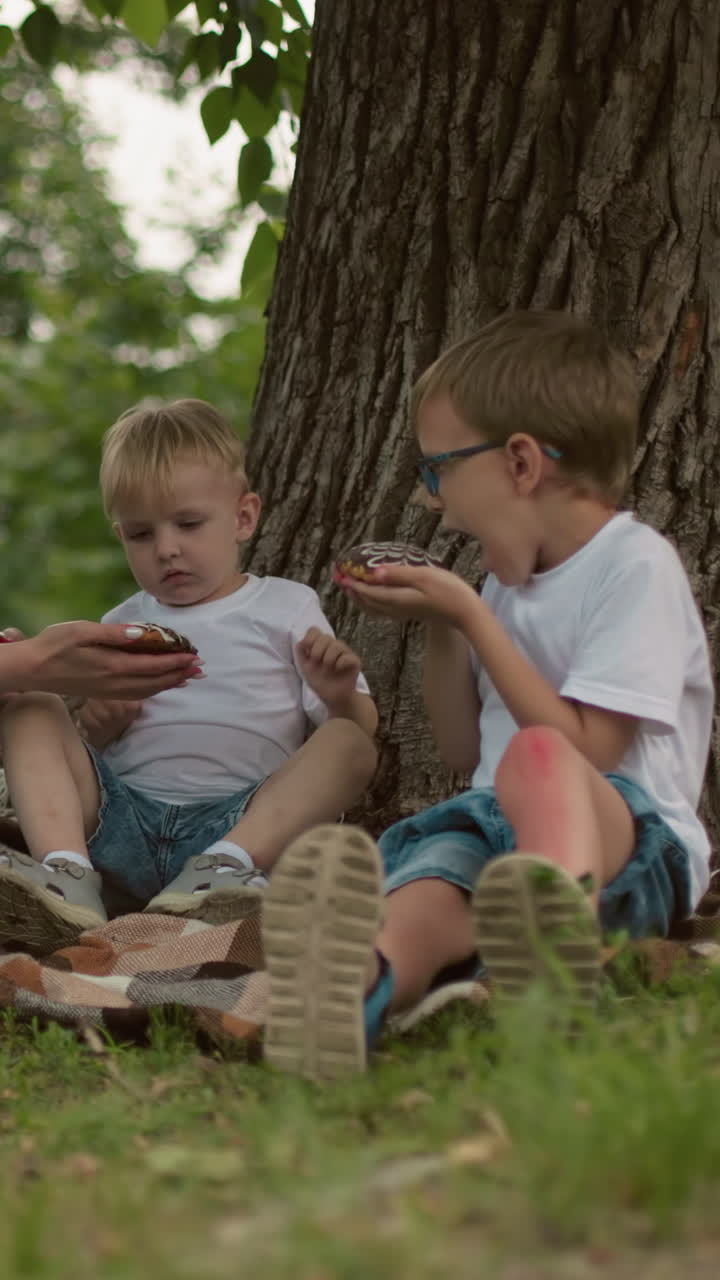 una madre y sus dos hijos se sientan en el suelo bajo un árbol, la madre ofrece un bocadillo a su hijo menor, quien lo rechaza, señalando hacia el bocadillo del hermano mayor