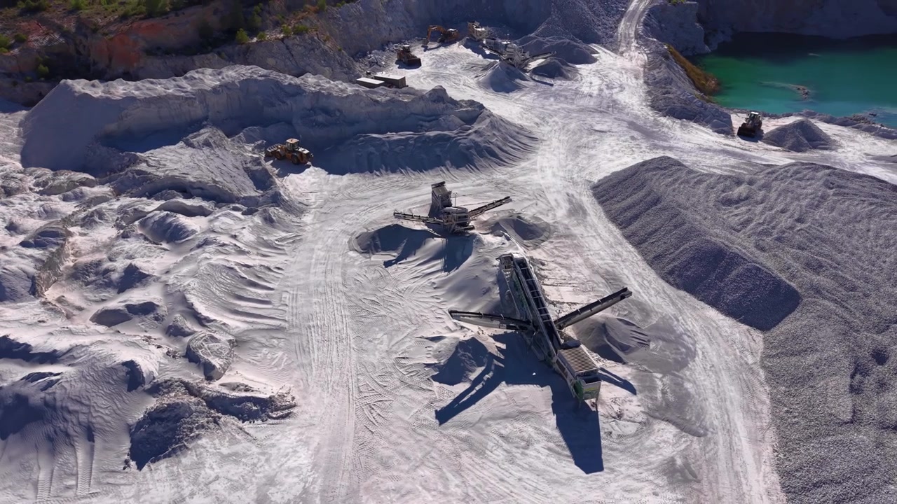 Mobile screening machines operating in a white gypsum open pit quarry, with mineral piles, heavy equipment and a turquoise water reservoir visible from above