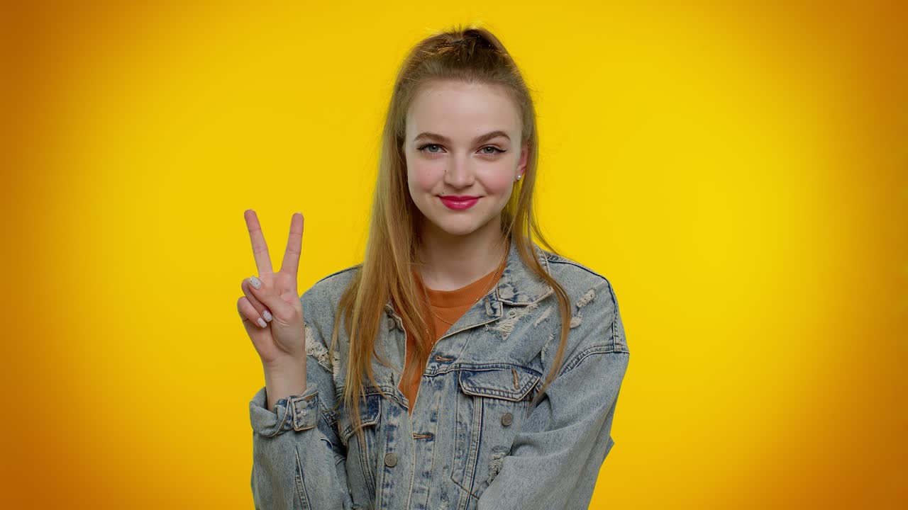 아름답고 세련된 미소 짓는 소녀는 승리의 표시를 보여주고, 성공과 승리를 희망하며, 평화의 몸짓을 합니다.