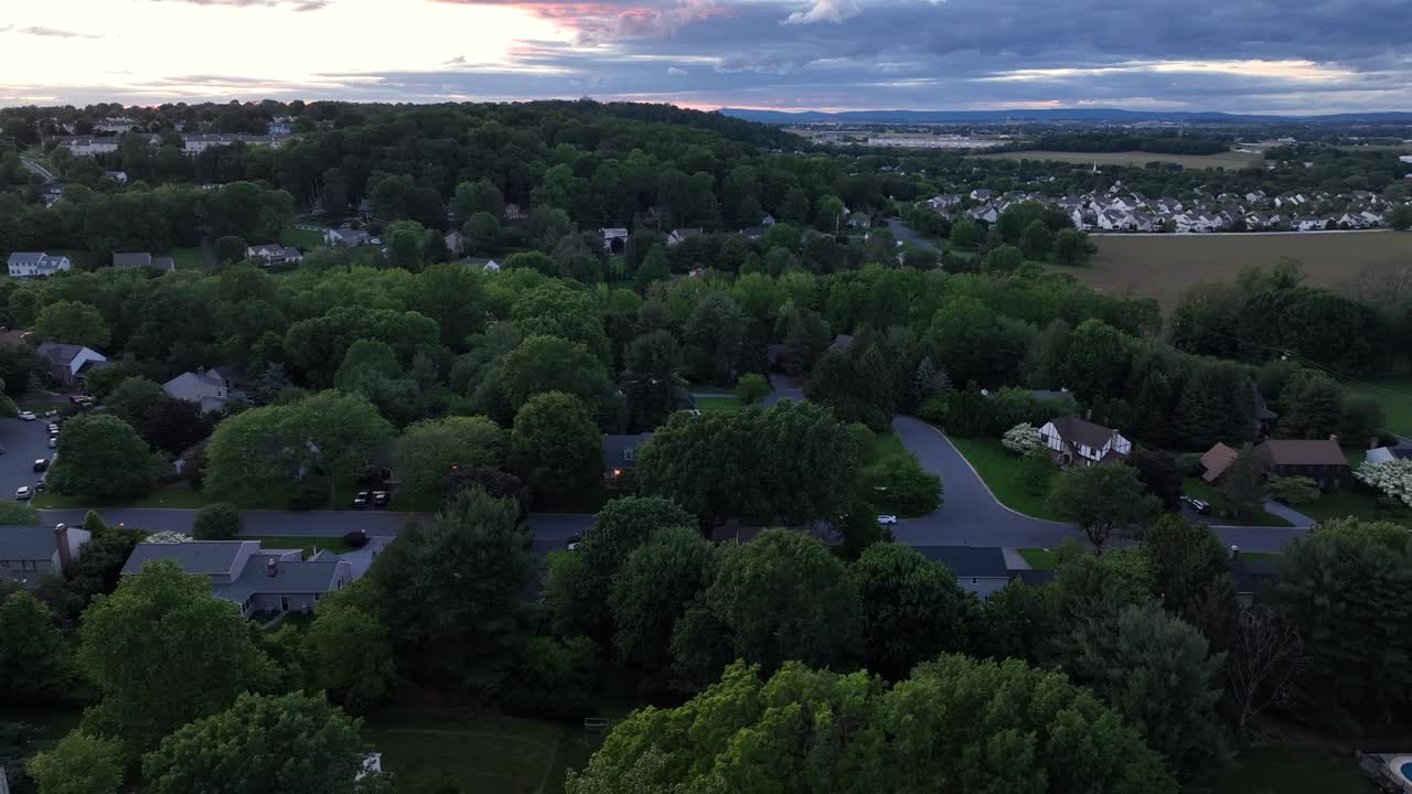 Grey roods of houses and homes between green trees in USA. Aerial lateral wide shot. Dusk scene after golden sunset. Clouds at sky. Quiet calm suburb district. Pennsylvania, United States.