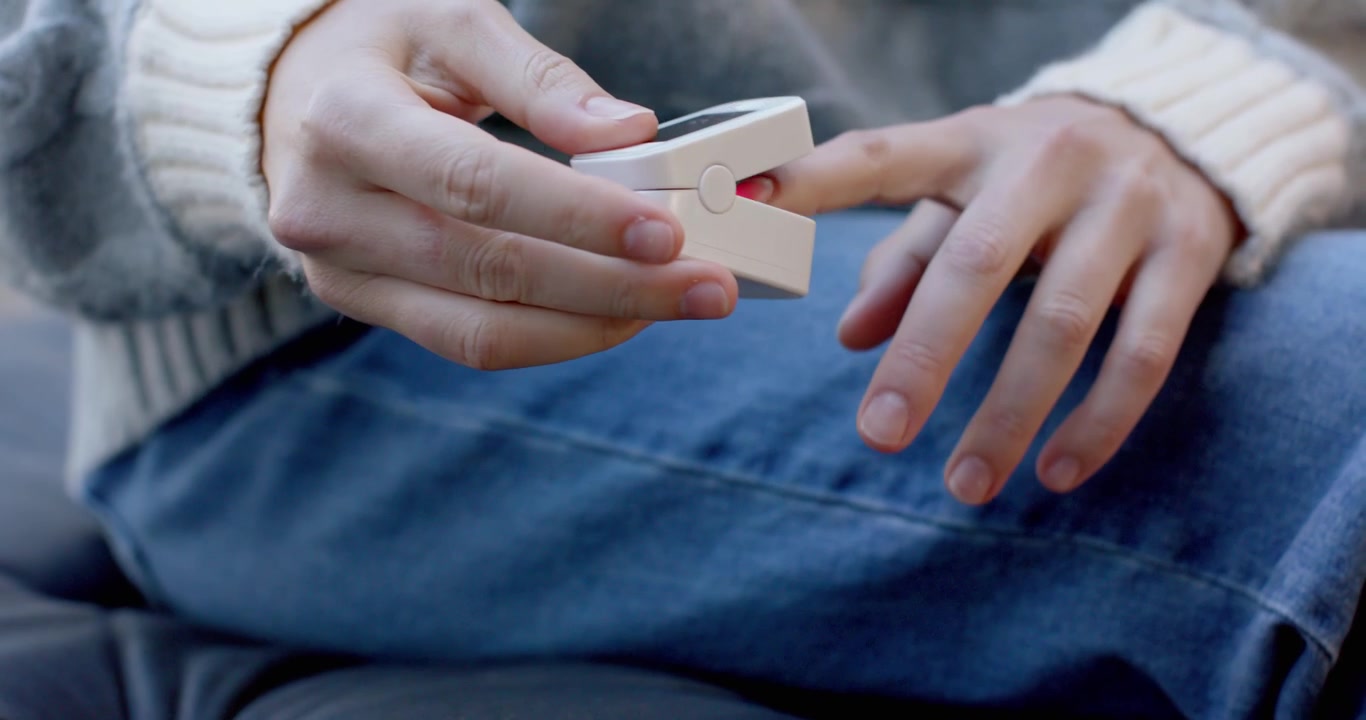 Woman in sweater and jeans tapping pulse oximeter on couch, clipping on finger, checking oxygen