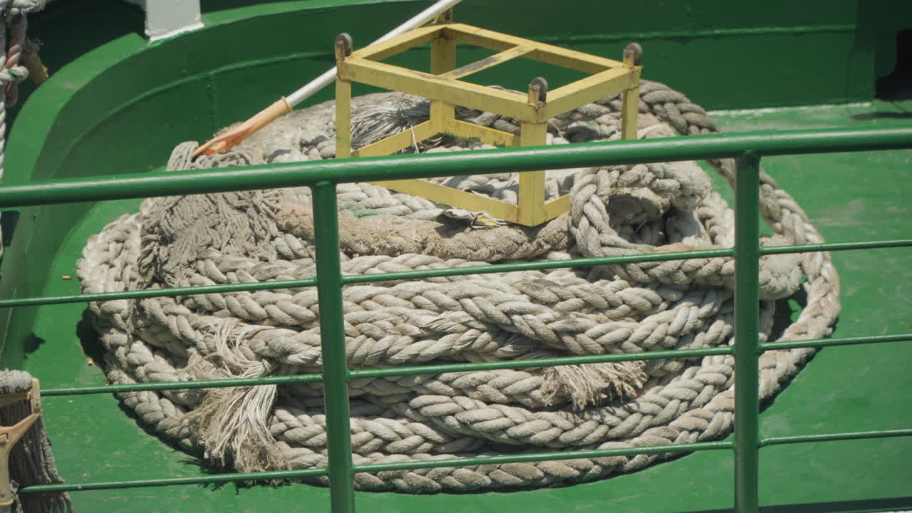Primo piano di corda avvolta vicino a ringhiera verde, spessa cima intrecciata avvolta all'interno del pozzetto del ponte con cuneo di stivaggio giallo, fibre spruzzate di sale, dettaglio di trama tattile, scena marittima pronta per l'ispezione, stock ad alto dettaglio.