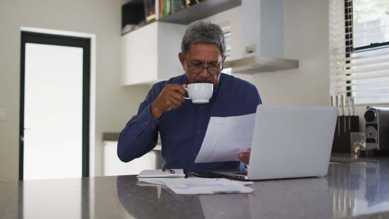 Uomo anziano multietnico in cucina che usa il portatile e beve caffè.