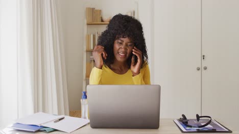 Afroamerikanische-Frau-Benutzt-Laptop-Und-Spricht-Auf-Dem-Smartphone,-Während-Sie-Zu-Hause-Auf-Ihrem-Schreibtisch-Sitzt