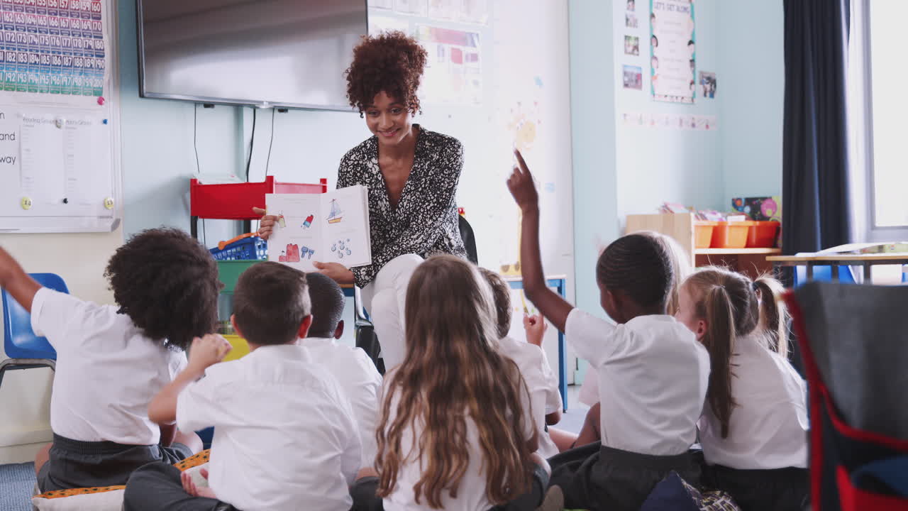 Alunno della scuola primaria in uniforme alza la mano per rispondere a una domanda mentre un'insegnante legge un libro