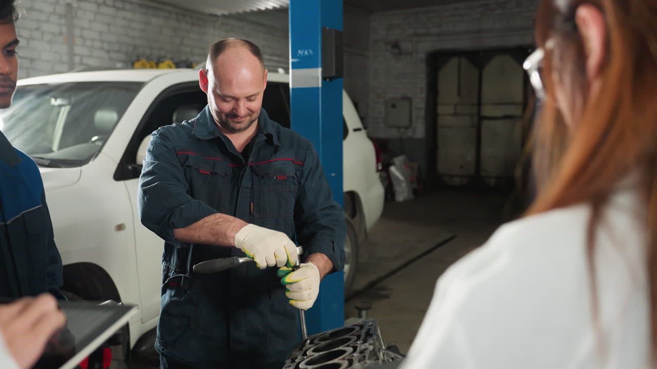 los estudiantes observan a un mecánico que demuestra el uso de llaves en el motor de un automóvil en un taller mecánico, el mecánico sonríe mientras maneja la herramienta, un espacio de trabajo industrial lleno de vehículos
