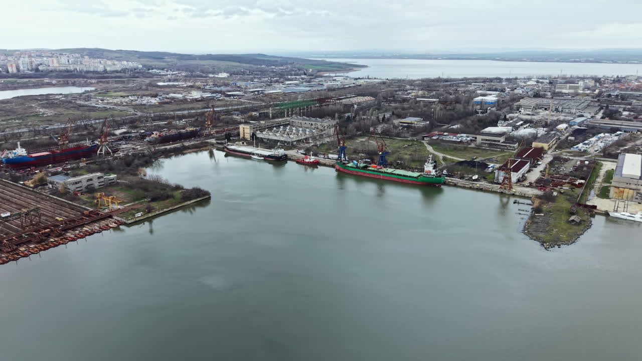 Navi cargo attraccano in un porto vicino a una città con edifici e un fiume durante una giornata nuvolosa