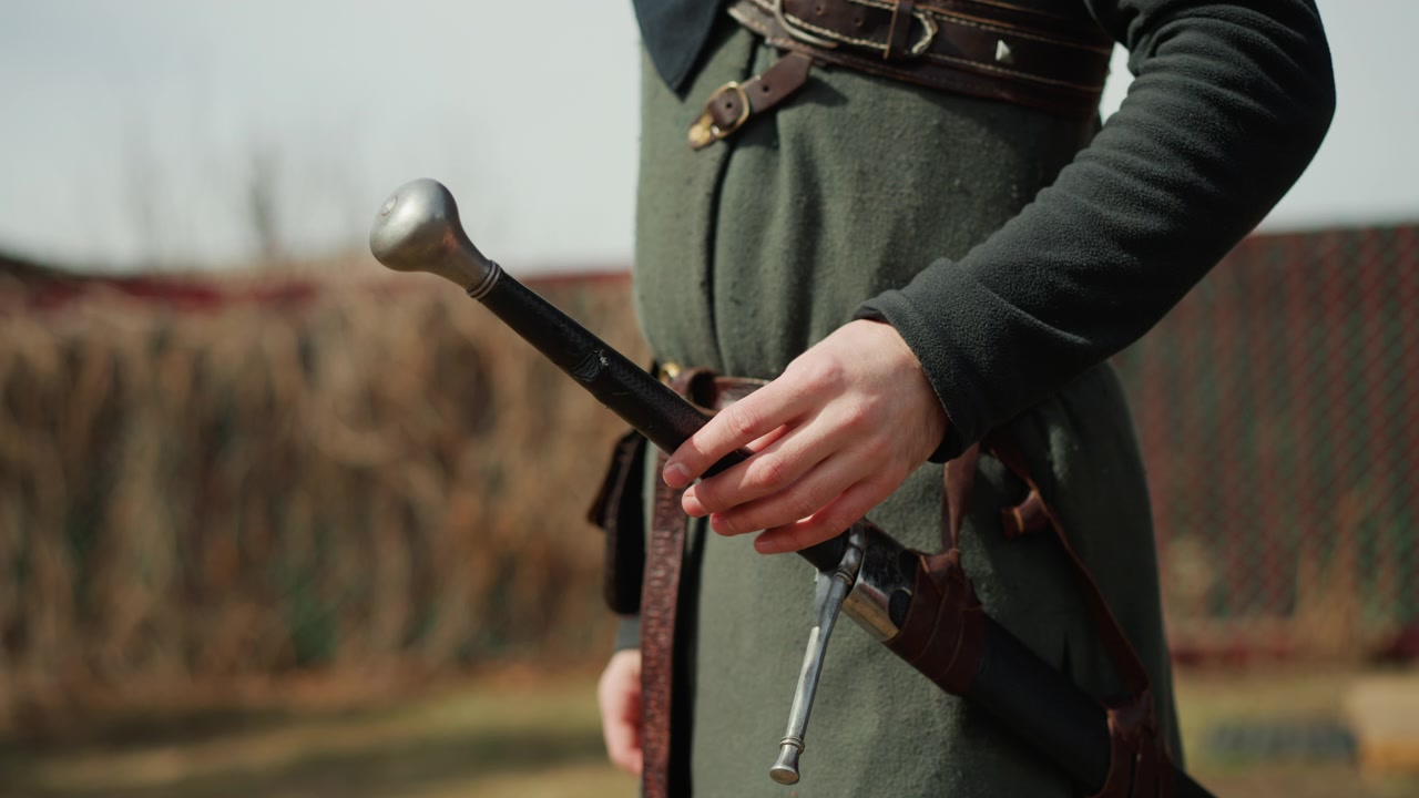 Vista a mezzo busto di una persona con una tunica in stile medievale che estrae parzialmente la spada dal fodero con una mano mentre stabilizza il fodero con l'altra, mostrando movimento controllato, posizionamento delle dita, dettagli in pelle e