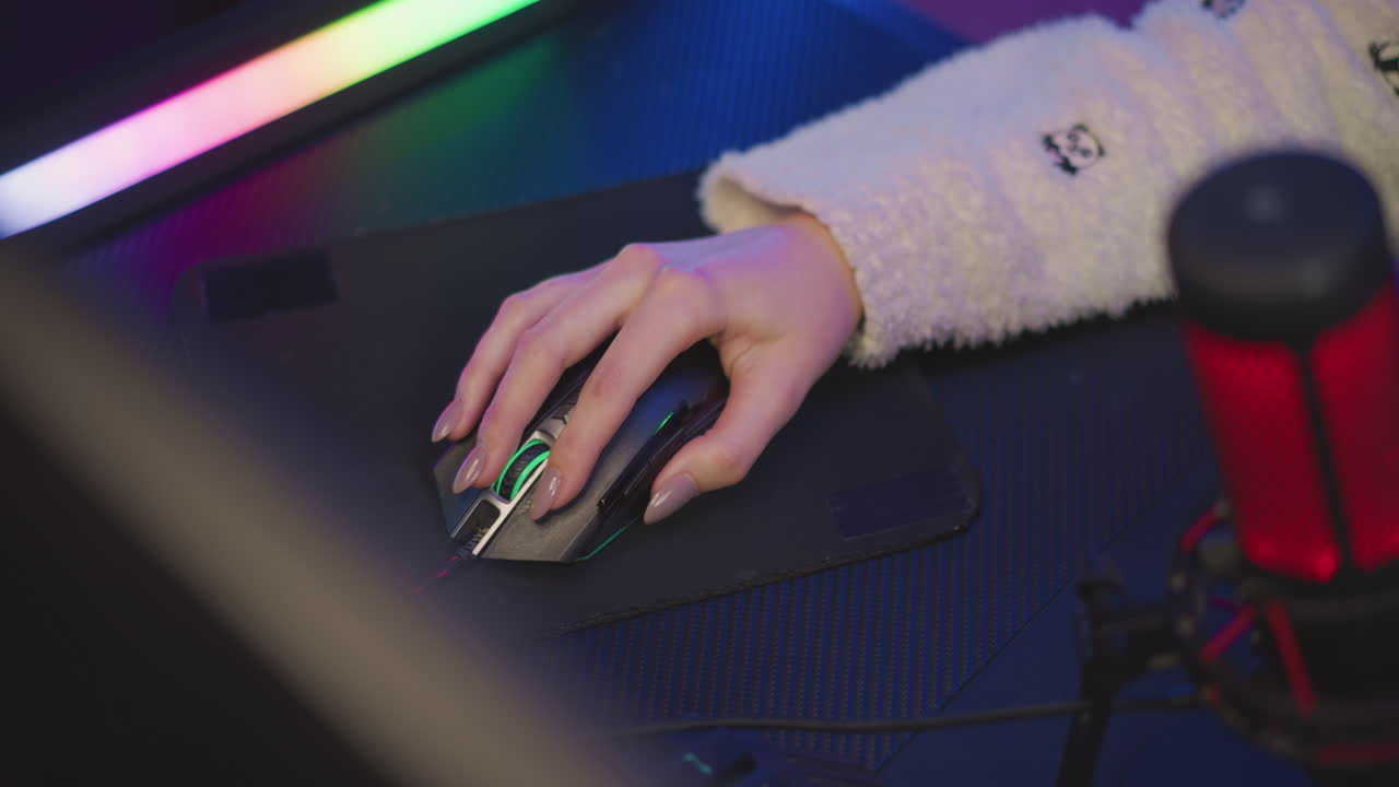 Aerial view of lady hand with polished nail in fluffy sweater featuring teddy bear design using illuminated gaming mouse on black pad, surrounded by colorful ambient lighting and streaming microphone