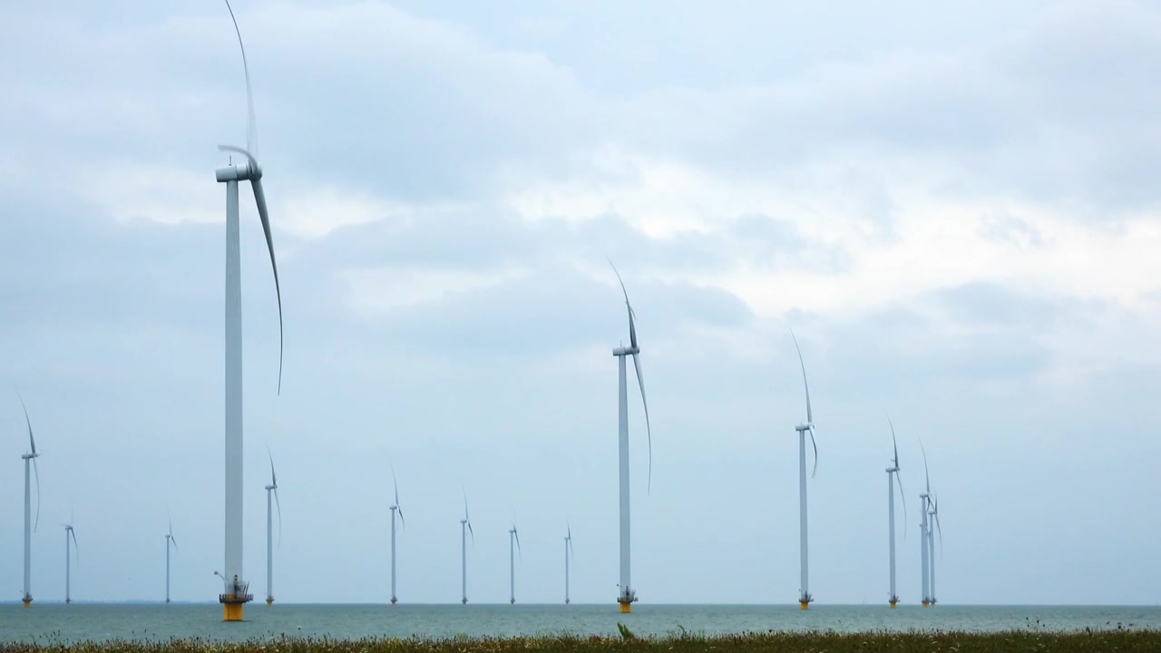 Ampio paesaggio costiero che mostra turbine eoliche offshore che si ergono su acque marine calme sotto un cielo nuvoloso drammatico e infrastrutture rinnovabili.