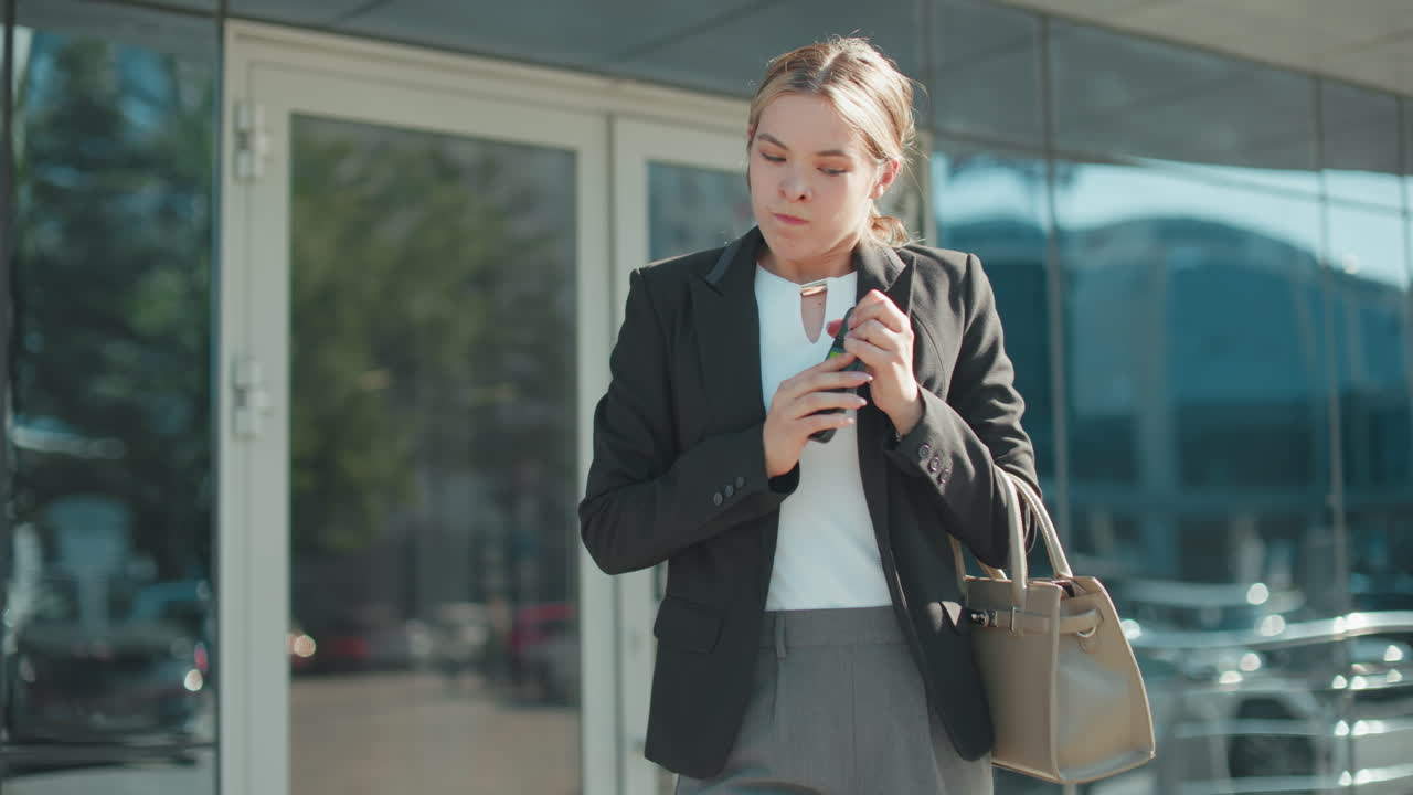 Furious lady on phone drops device in frustration while continuing call, exiting building in professional outfit, holding handbag, urban glass structure reflects cars and cityscape